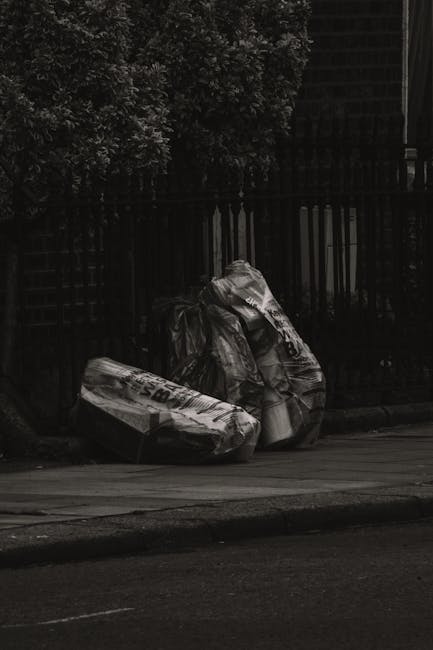 A black wheeled rubbish bin positioned on a quiet urban sidewalk during nighttime, with the lid partially open revealing a variety of discarded materials including paper, cardboard, and plastic waste.