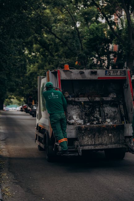 A sanitation worker wearing a high-visibility yellow vest and dark clothing operates a waste collection vehicle, which is primarily white with red and orange reflective markings. The worker is standin