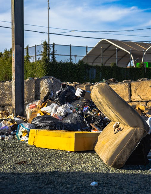 Close-up view of a pile of mixed rubbish and waste materials situated on a gravel surface, including black plastic garbage bags, a yellow plastic container, and a worn, discolored brown sofa armrest w